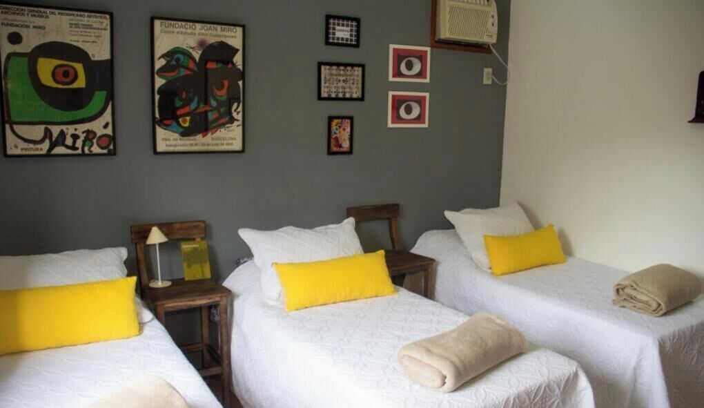A hotel room with three beds, each with a white bedspread and a yellow pillow. There are framed artworks on the wall, a small table with a lamp, and a window with a white curtain.