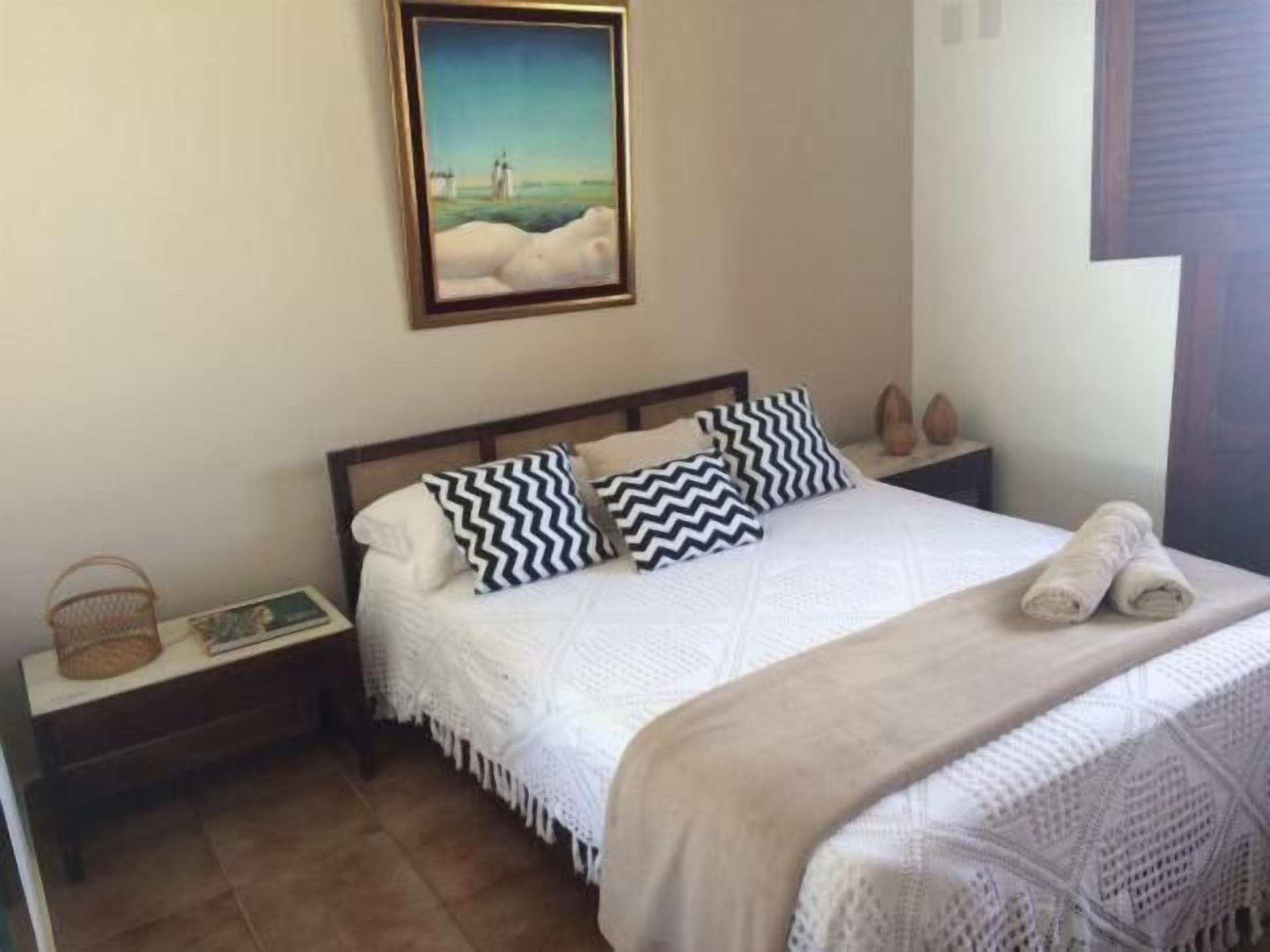 A neatly made bed with white linens and two black and white patterned pillows, a small bedside table with a book and a basket, and a framed painting above the bed.