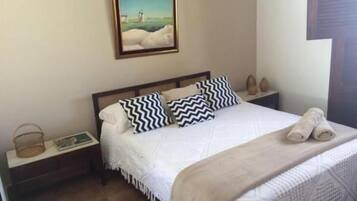 A neatly made bed with white linens and two black and white patterned pillows, a small bedside table with a book and a basket, and a framed painting above the bed.