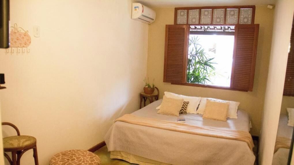 Un dormitorio con una cama, una ventana con contraventanas, una mesita con una planta y una silla.