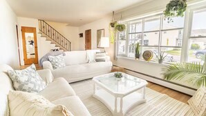 Ein helles Wohnzimmer mit einem weißen Sofa, einem Glastisch und einem großen Fenster mit Pflanzen.
