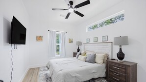 Un dormitorio con cama, ventilador de techo, televisor, ventana con cortinas y mesita de noche con lámpara.