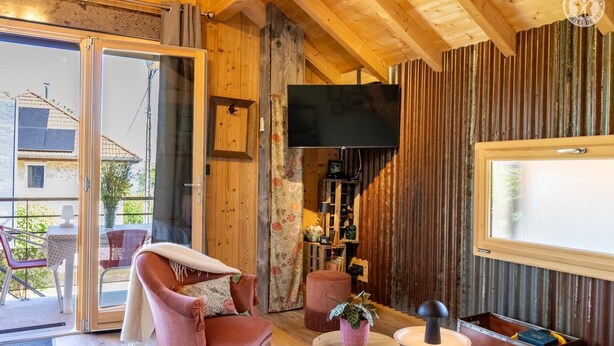 Una habitación acogedora con techo de madera, un televisor montado en la pared, un sillón cómodo, una mesita pequeña y una ventana con cortinas.