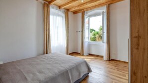 Un dormitorio con cama, piso de madera, ventana con cortinas y vistas a la vegetación.