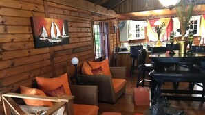 Interior acolhedor de uma cabana, com móveis de vime, uma pintura de barcos à vela e uma sala de jantar com mesa e cadeiras.