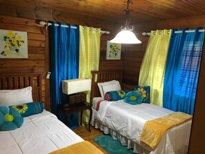 A cozy room with two single beds, wooden headboards, a nightstand, a lamp, and colorful curtains.