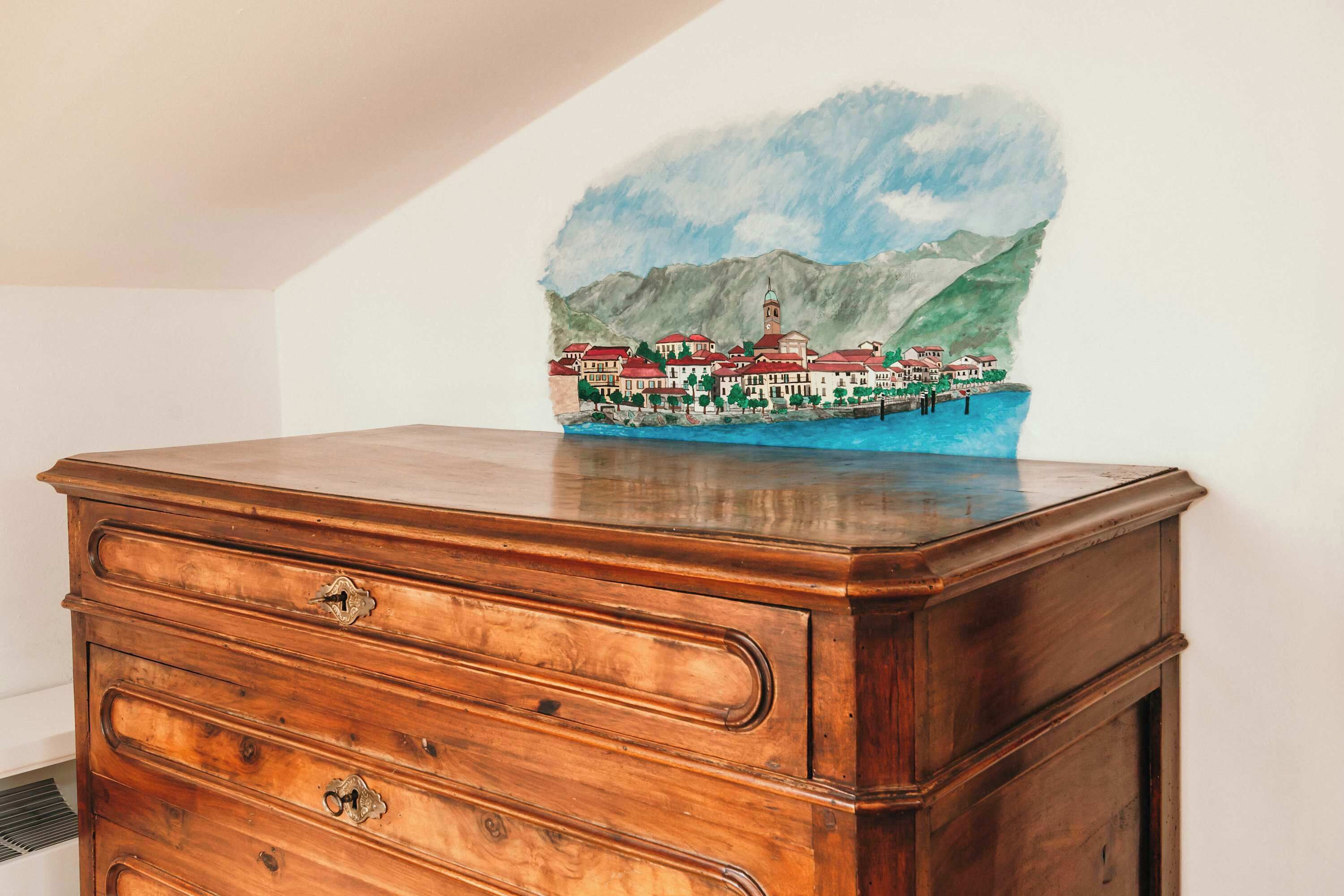 A wooden dresser with a drawer, a small painting of a coastal town, and a vent on the wall.
