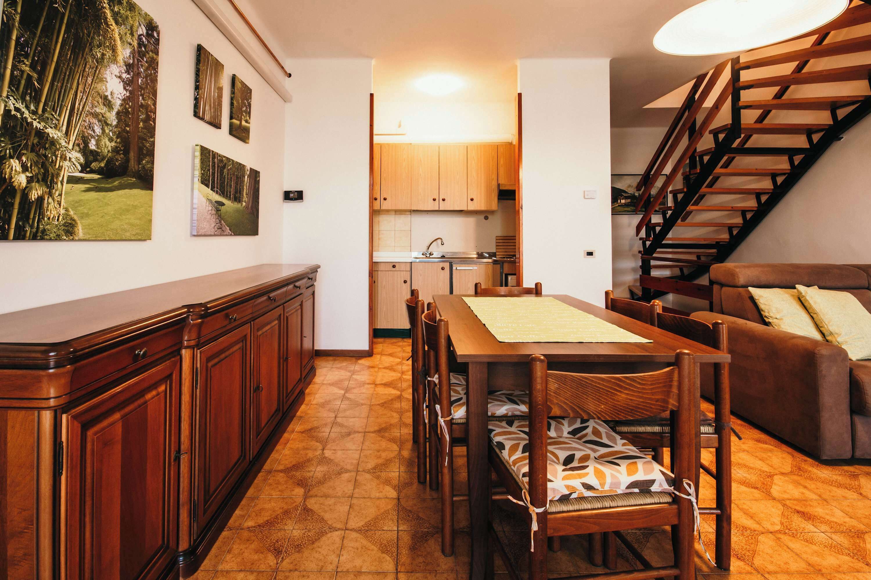 A dining area with a wooden table and chairs, a kitchen in the background, and a staircase.