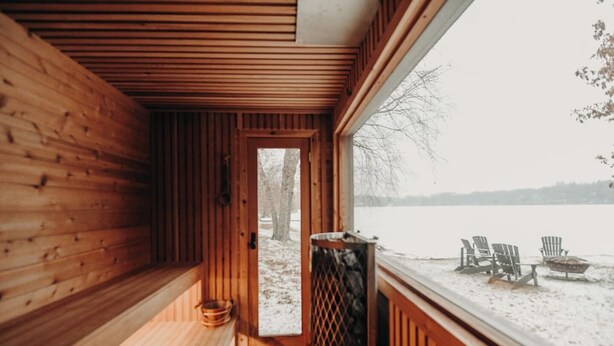 A wooden sauna with a bench and a window leading to a snowy outdoor area with chairs. - LAKESIDE CITY GETAWAY (Roseville)