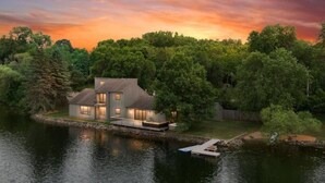 A modern house by a lake with a dock, surrounded by dense trees and a sunset sky. - LAKESIDE CITY GETAWAY (Roseville)