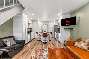 A modern kitchen with white cabinets, a dining area with a wooden table and bar stools, a black leather sofa, and a TV mounted on the wall.