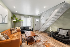 Un salon moderne avec un canapé en cuir noir, un divan en cuir marron et une table basse en bois. Un escalier se trouve à droite et une porte, au fond.