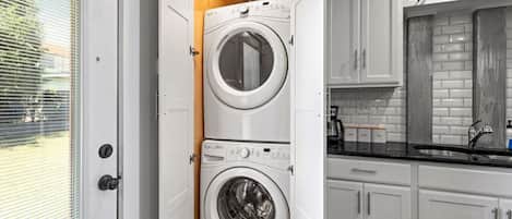 A laundry room with two stacked front-loading washing machines and a kitchen area with white cabinets and a black countertop.