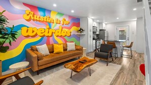A modern living room with a colorful mural, a brown sofa, a wooden coffee table, and a dining area with chairs. - Retro SoCo GetawayTownhome! (Austin)