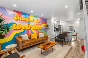A modern living room with a colorful mural, a brown sofa, a wooden coffee table, and a dining area with chairs. - Retro SoCo GetawayTownhome! (Austin)