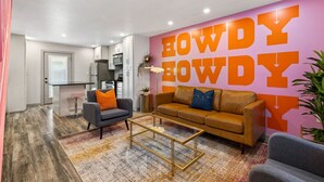A modern living room with a large wall mural, a sofa, a coffee table, and a kitchen in the background. - Pink Palace SoCo Dream Townhome! (Austin)