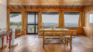 Et rum med træpaneler, et bord og stole, store vinduer med gardiner og udsigt til landskabet.