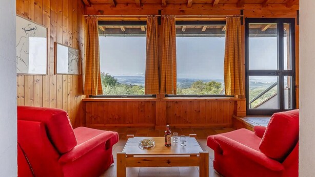 Et værelse med trævægge og -loft, to røde stole, et bord med en flaske og glas, samt udsigt over landskabet gennem store vinduer.