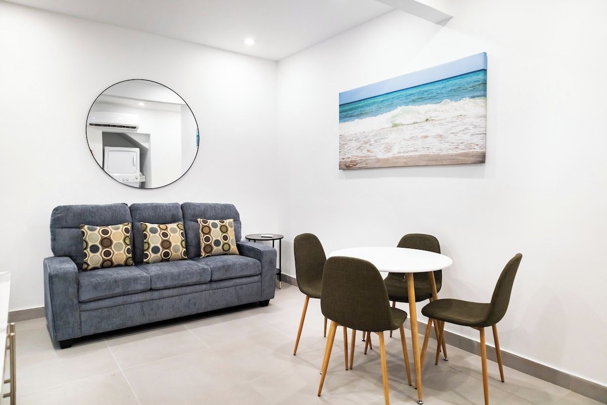 Un salon moderne avec un canapé gris, un miroir rond, un tableau représentant une plage et un coin repas avec une table et des chaises blanches.