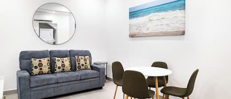 Una sala moderna con un sofá gris, un espejo redondo, un cuadro de playa y un comedor con una mesa y sillas blancas.