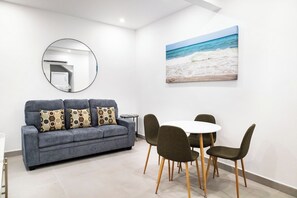 Ein modernes Wohnzimmer mit einem grauen Sofa, einem runden Spiegel, einem Strandbild und einem Essbereich mit einem weißen Tisch und Stühlen.