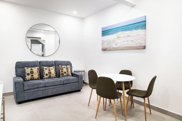 A modern living room with a grey sofa, a round mirror, a painting of a beach, and a dining area with a white table and chairs.