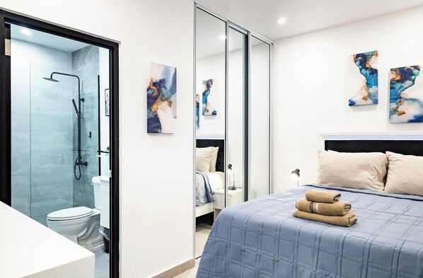 A modern bathroom with a glass shower enclosure, a toilet, and a bed with a blue quilted bedspread and beige pillows.