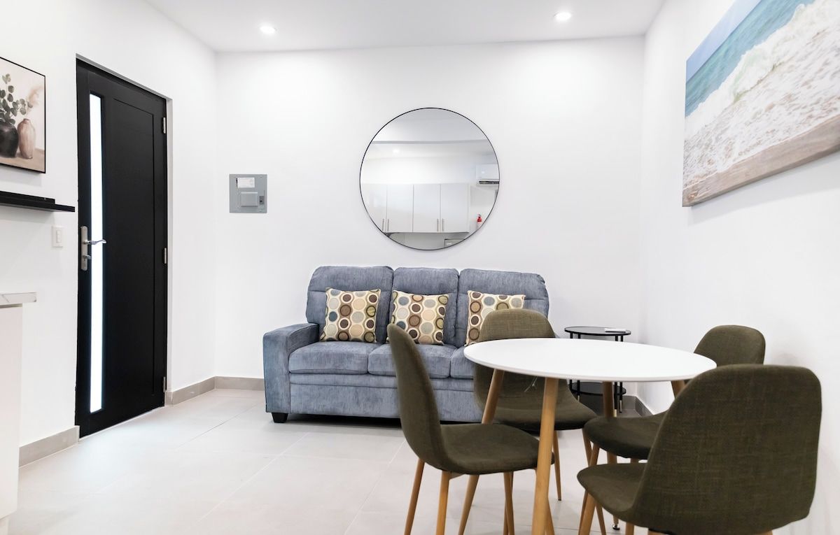 A modern living room with a grey sofa, patterned cushions, a round mirror, and a round dining table with chairs.