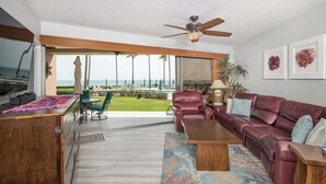 A spacious living room with a leather sofa, a wooden coffee table, and a TV on a wooden cabinet. A large window offers a view of the ocean and a patio area with outdoor furniture.