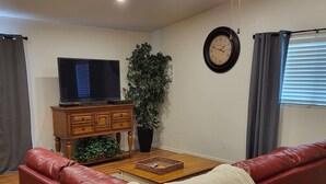 A living room with a red leather sofa, a wooden coffee table, a television on a wooden stand, a ceiling fan, a clock, and a window with curtains. - Charming 2BD / 1 BA farmhouse on 2.5 acres w/mountain views in fabulous Glendale (Glendale)