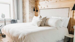 Un dormitorio con cabecera de madera, pared de ladrillo, cama con ropa de cama blanca, una mesita de noche y una pequeña mesa con lámpara.