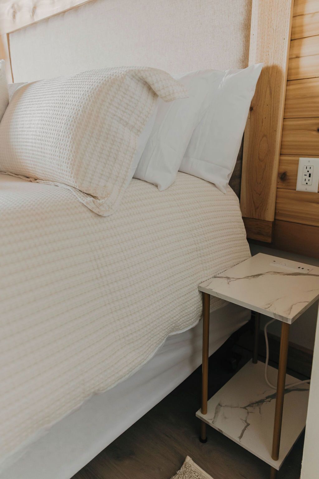 A bed with a white quilted cover and two pillows, a small bedside table with a marble-like surface, and a wooden wall panel with a power outlet.