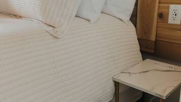 A bed with a white quilted cover and two pillows, a small bedside table with a marble-like surface, and a wooden wall panel with a power outlet.