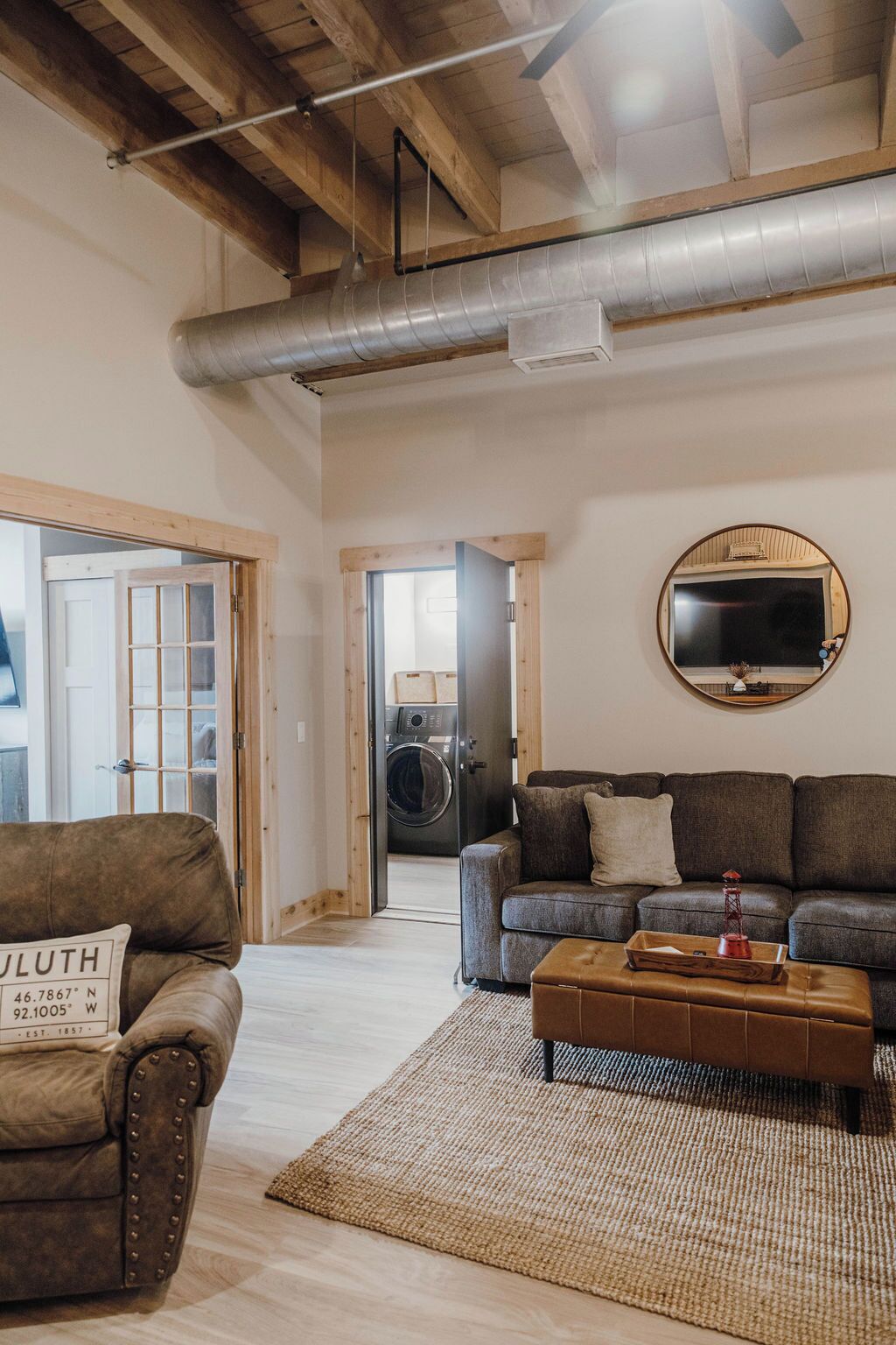 Un salon spacieux avec un grand canapé, une table basse et un téléviseur fixé au mur. Une porte en bois donne accès à une autre pièce, et on peut apercevoir un lave-linge à travers l’ouverture de la porte.