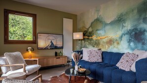 Een woonkamer met een blauwe bank, een houten salontafel, een witte fauteuil en een groot abstract schilderij aan de muur.