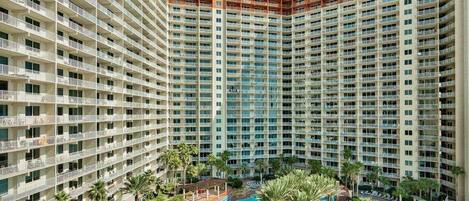 A high-rise hotel with multiple balconies, a central swimming pool, and surrounding palm trees.