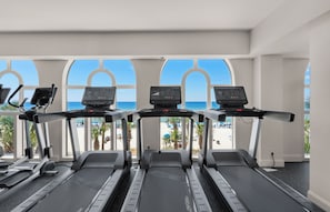 Academia com esteiras e equipamentos de ginástica, janelas amplas com vista para a praia.