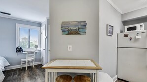 A compact kitchen with a wooden table, two stools, a refrigerator, and a painting of a dock. - Cozy Holiday Hideaway, Sleeps 4! Steps to the Beach! Renovated (Panama City Beach)