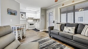 A modern living room with a gray sofa, a wooden ceiling fan, and a kitchen in the background. - Cozy Holiday Hideaway, Sleeps 4! Steps to the Beach! Renovated (Panama City Beach)