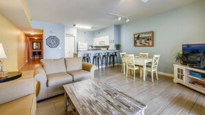 Een woonkamer met een bank, een salontafel en een eethoek met stoelen en een tafel. Achtergrond is een keuken met een magnetron en kastjes.