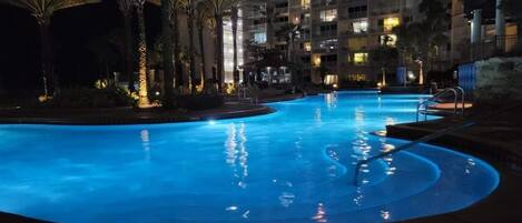 A nighttime swimming pool with palm trees and a high-rise building in the background.
