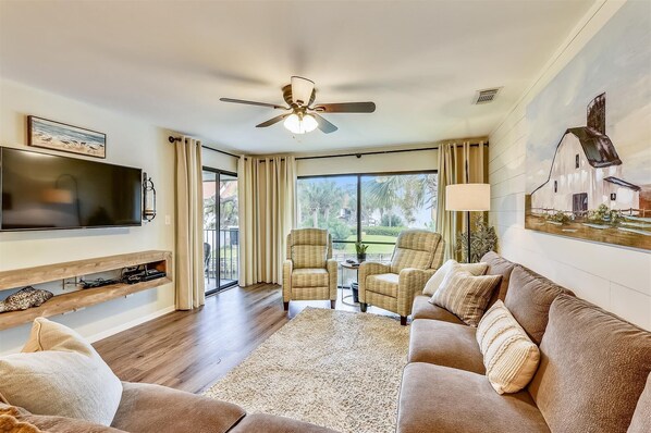 A spacious living room with a flat-screen TV, comfortable seating, a ceiling fan, and a large window offering a view of the outdoors. - Edgewater Villas #2108 - 2 Bedroom/2Bath Sleeps 8.  Free Fun! (Panama City Beach)