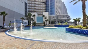 Zona de piscina de hotel con una fuente, palmeras y un edificio moderno al fondo.