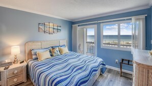 Ein Schlafzimmer mit Bett, Nachttisch und Blick auf den Strand durch ein Fenster.