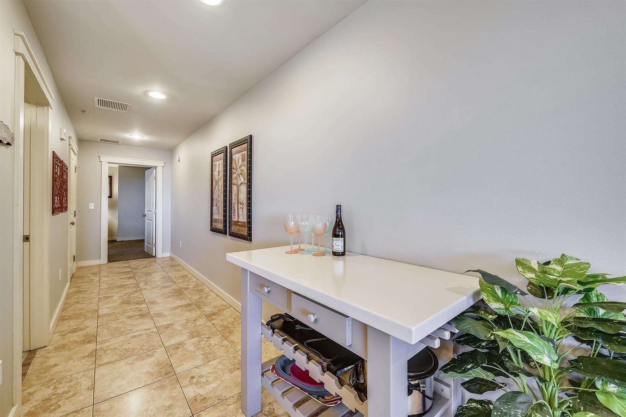 Un pasillo con una mesa blanca que sostiene una botella, vasos, una planta y una puerta que da a otra habitación.
