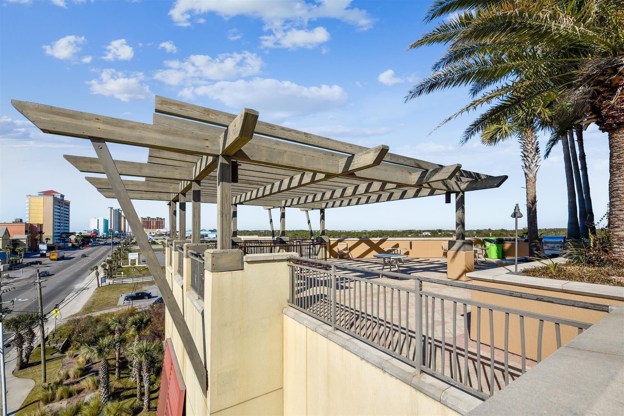 A covered outdoor area with a pergola structure, a paved walkway, and a view of a cityscape with buildings and palm trees.
