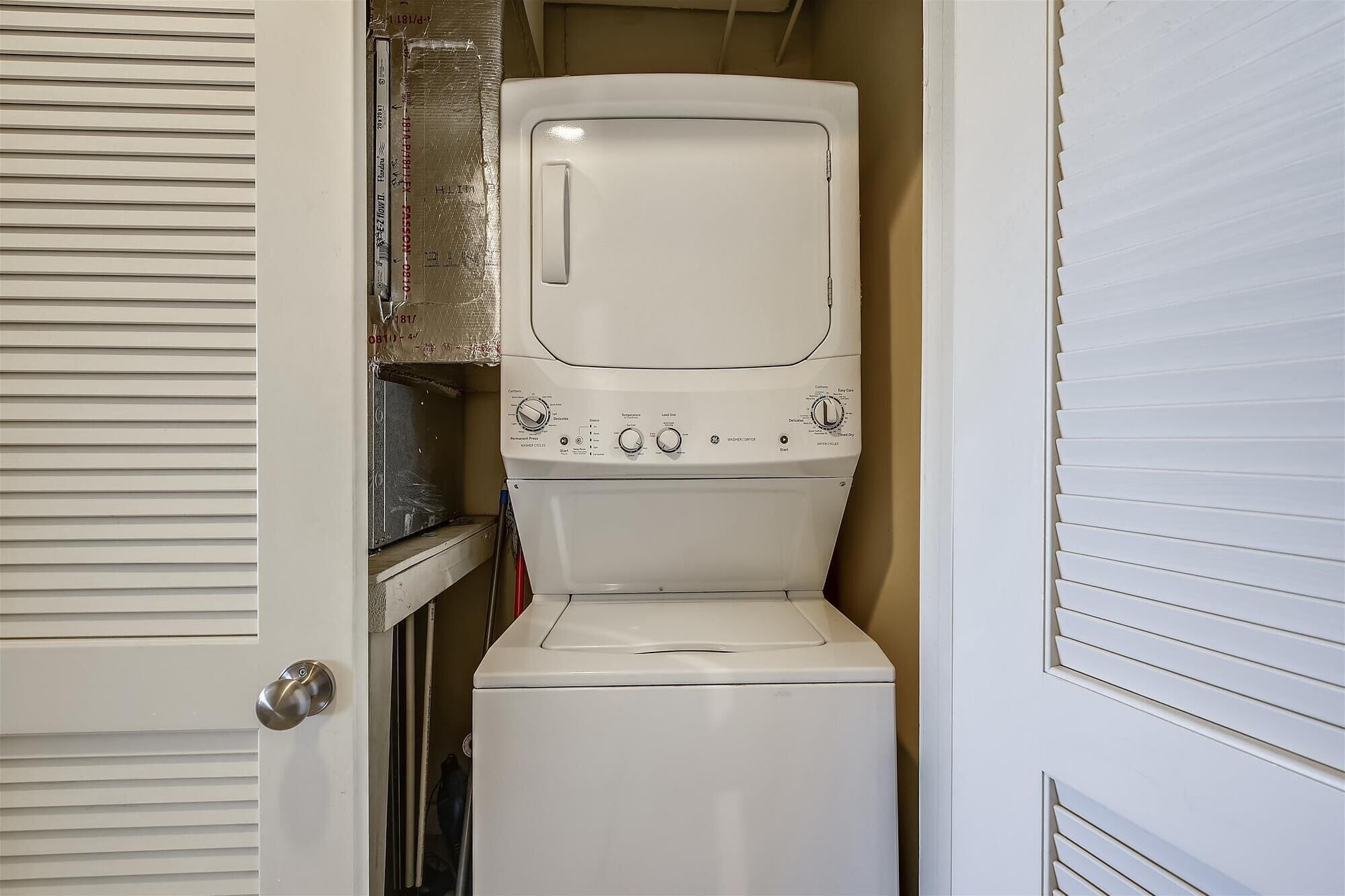 Une petite buanderie équipée d’un lave-linge et d’un sèche-linge à chargement frontal, d’une petite étagère et d’une porte dotée de panneaux à persiennes horizontales.