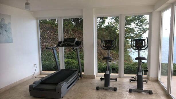 A spacious room with a treadmill, elliptical, and stationary bike, large windows with a view of greenery, and a painting on the wall. - Romance Nest in Samana 7-202 (Samaná)