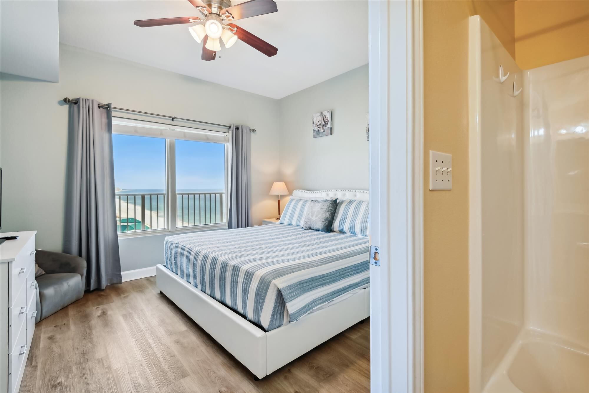 A bedroom with a bed, a desk, a chair, a window with curtains, a ceiling fan, and a view of the beach.