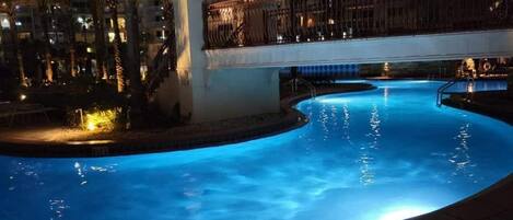 Una piscina nocturna de vora corbada, envoltada de palmeres i un edifici al fons.